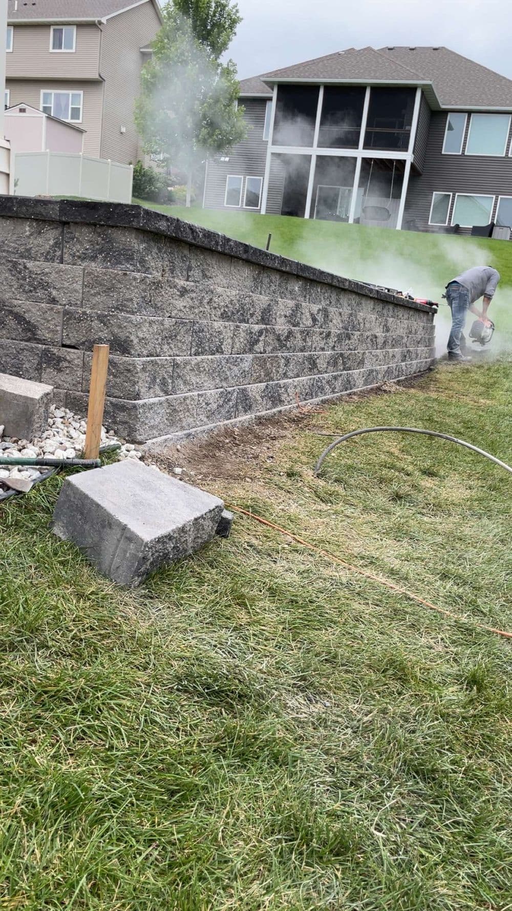 Man working on a stone retaining wall, surrounded by smoke and tools in a grassy yard.