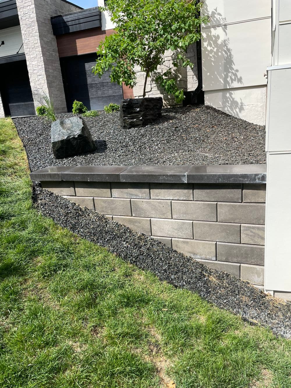 Diverse landscaping features with stone wall, gravel, and a tree near a modern home.