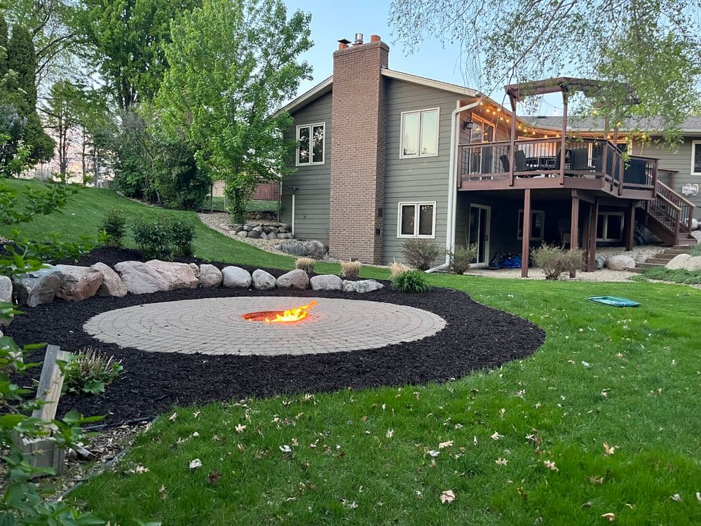 Backyard with fire pit surrounded by landscaping, deck, and lush green lawn.