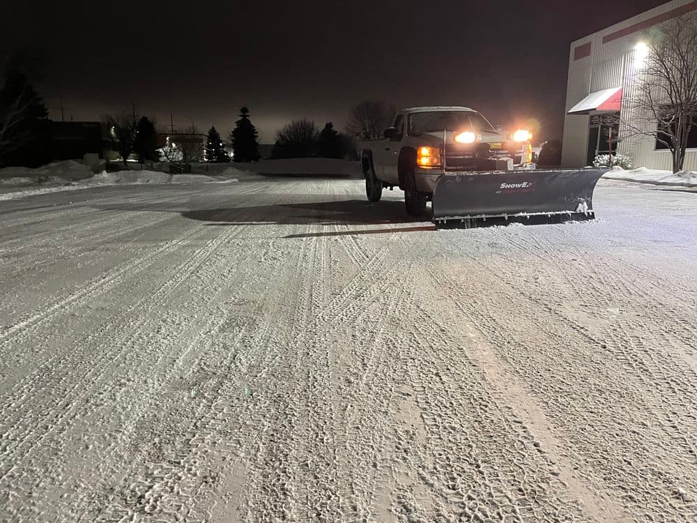 Snow plow truck clearing a snowy parking lot at night with ambient lighting.