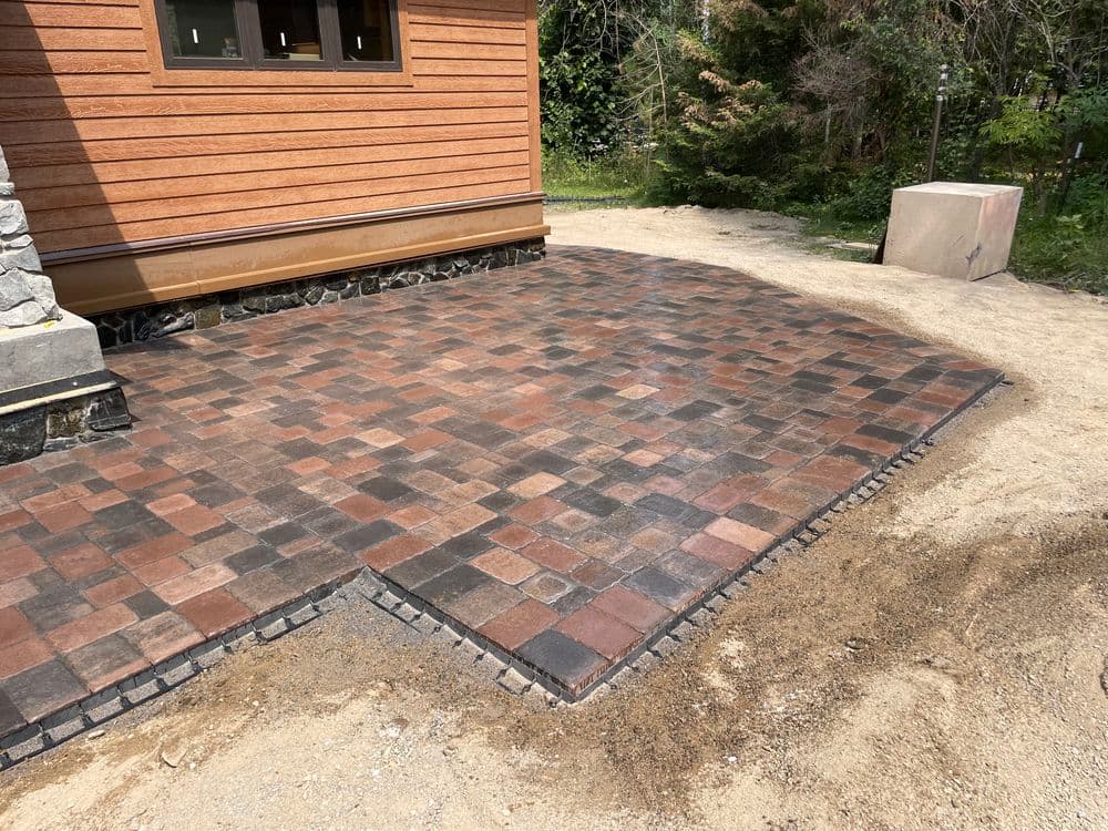 Paved patio area with interlocking stones near a wooden house and landscaped surroundings.