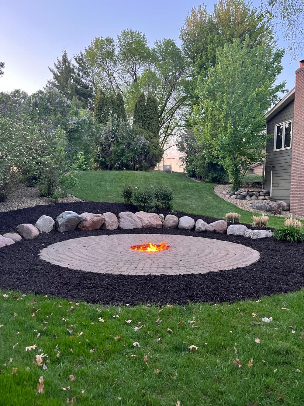 Stone fire pit surrounded by landscape features and greenery in a backyard setting.