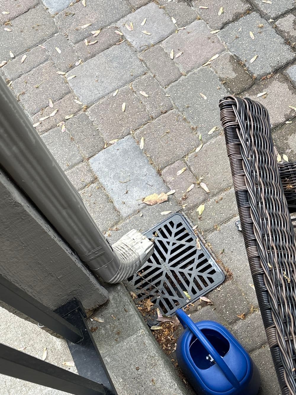 Drainage grate beside a porch with a blue watering can, surrounded by paved stones.